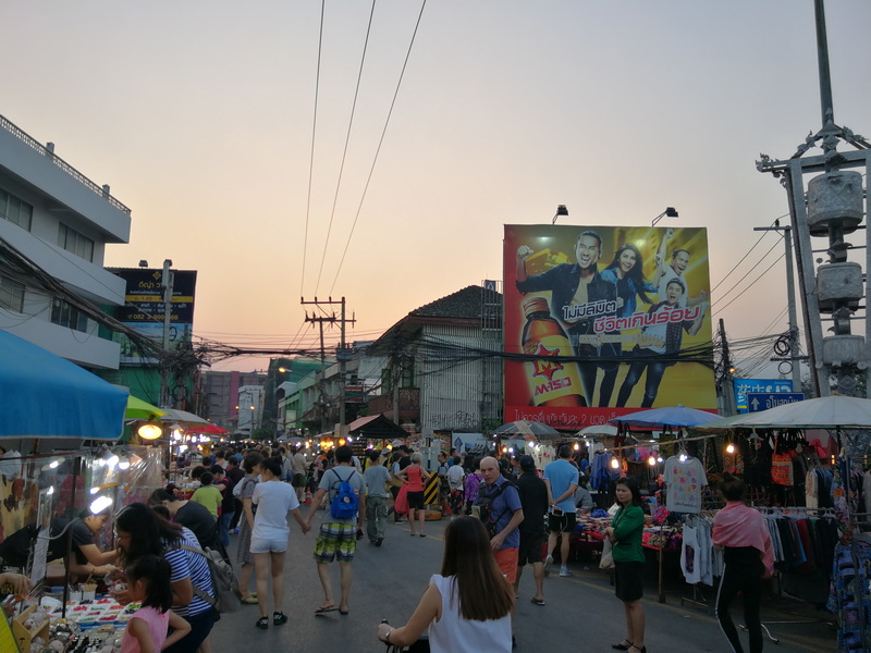 A lively street market filled with vendors and shoppers during sunset with a large billboard in the background at Wua Lai Walking Street Chiang Mai.