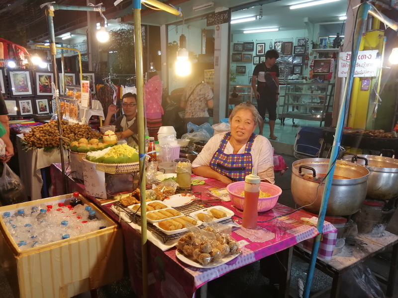 Elderly woman selling snacks at night market stall with people and vendor booths behind her at Saturday Walking Street Chiang Mai.