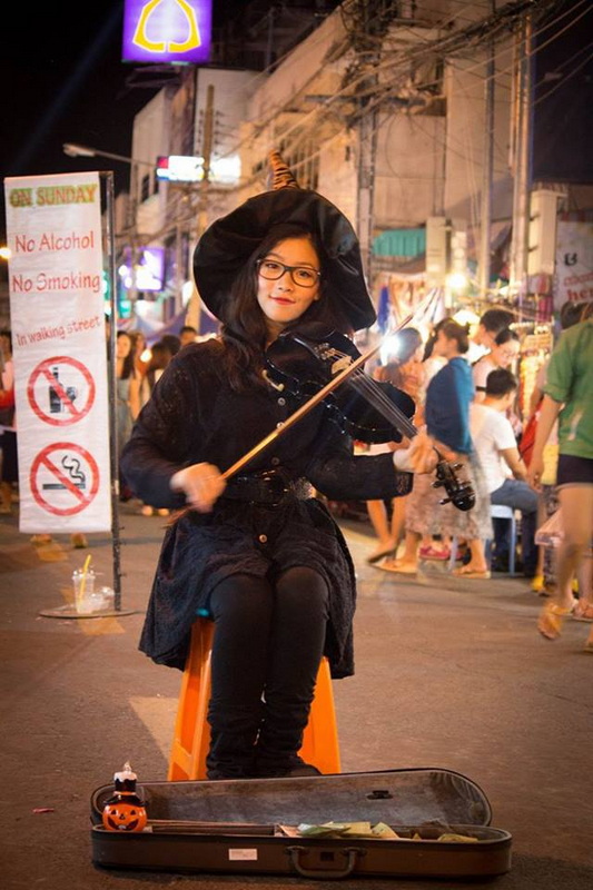 Night Market Musician at Sunday Night Market Chiang Mai Girl in witch costume playing violin at night market with crowd and lights at Sunday Night Market Chiang Mai.