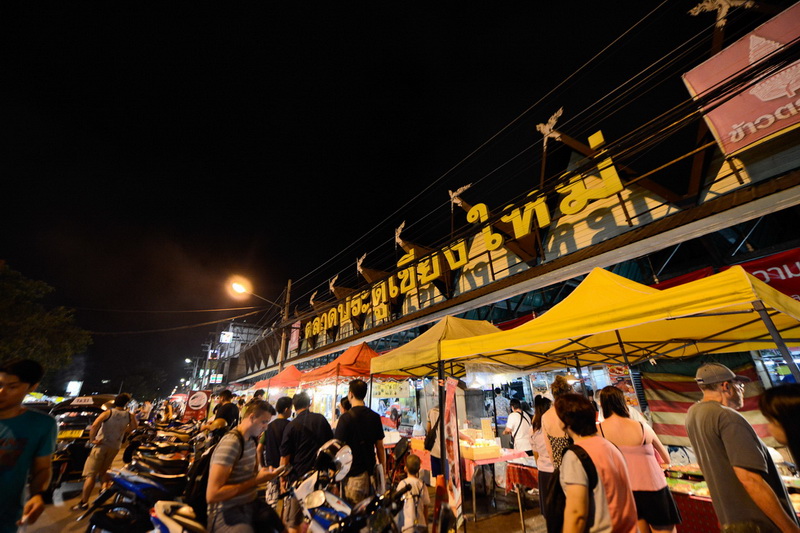 Night Market Scene at Best Night Markets In Chiang Mai Night market scene with stalls, tents, and visitors under a sign in Thai script at Best Night Markets In Chiang Mai.