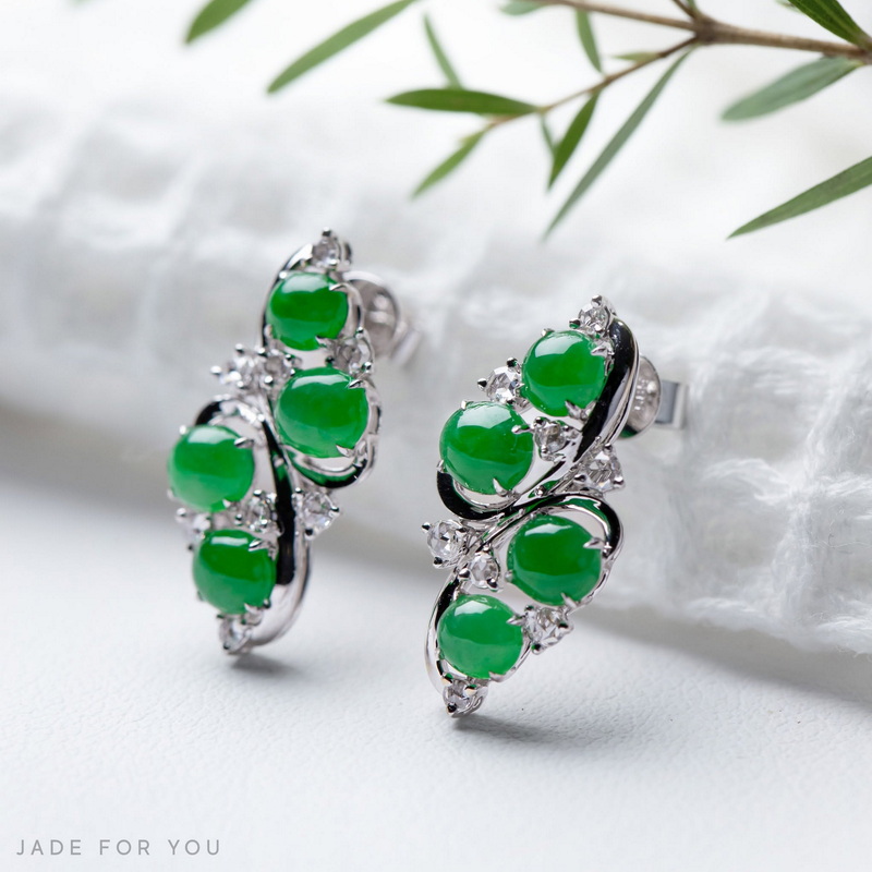 Silver earrings with green jade stones and diamonds on a white surface with green leaves behind at Chiang Mai Jade Factory.