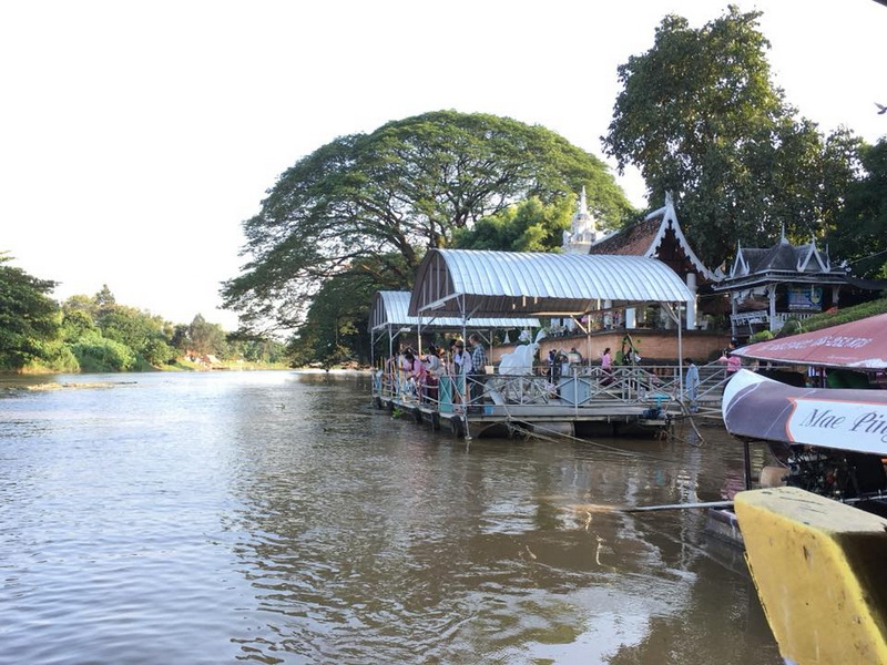 Boat with covered platform on river near a traditional Thai temple with trees at Chiang Mai Cruise To Wiang Kum Kam.