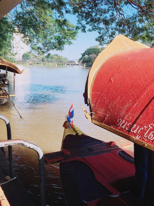 Boat docked by the river under trees with a bridge in the background at Chiang Mai Cruise To Wiang Kum Kam.