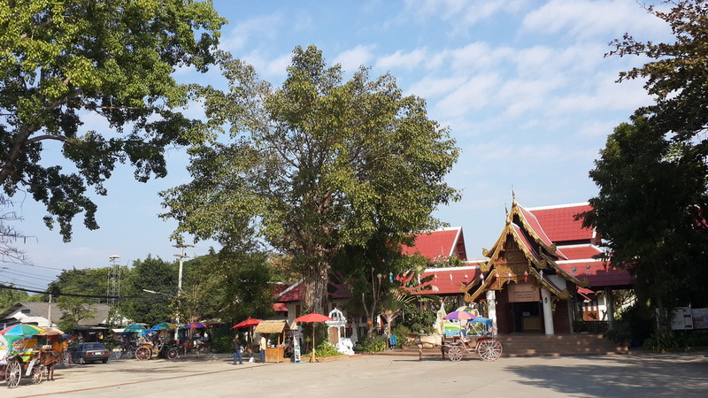 Thai temple with red roof and golden details, shaded by trees and busy street vendors at Chiang Mai Cruise To Wiang Kum Kam.