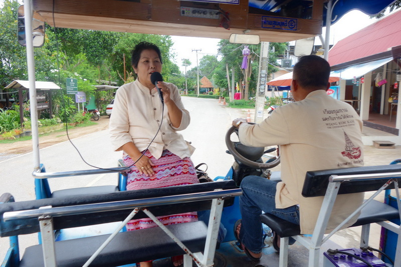 Woman speaking into a microphone while riding in a small vehicle on a village road at Chiang Mai Cruise To Wiang Kum Kam.