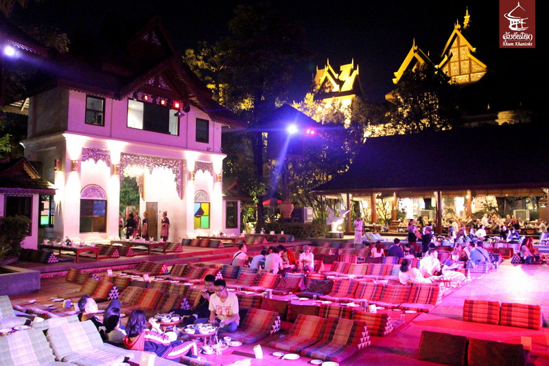 Night scene of outdoor dining with cushions, lights, and traditional Thai architecture in background at Khantoke Dinner.