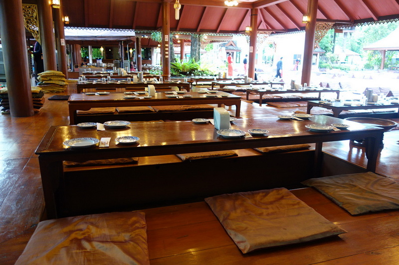 Interior of a traditional wooden dining space with low tables and cushions, overlooking a garden at Khum Khantoke.