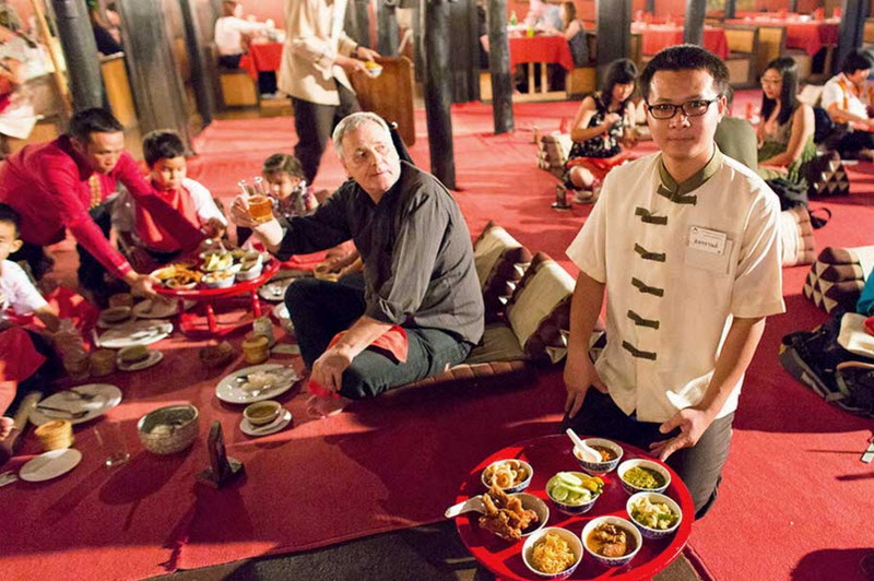 Indoor Dining Scene with Food at Old Chiangmai Cultural Center People seated on a red carpet with food bowls, some children in the background enjoying a meal at Old Chiangmai Cultural Center.
