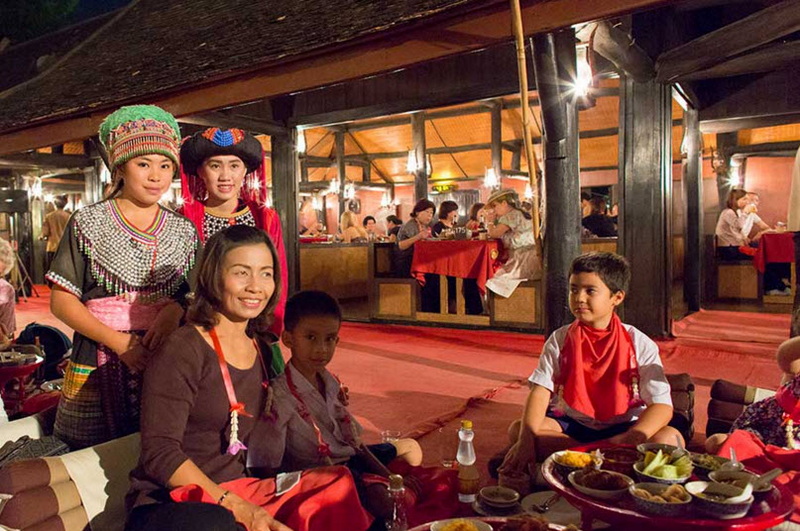 Traditional Cultural Dining Scene at Old Chiangmai Cultural Center People wearing traditional costumes dining inside a wooden building with food supplies at Old Chiangmai Cultural Center.