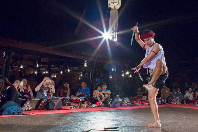 Male performer spinning sword during a dance in front of seated audience indoors at Old Chiangmai Cultural Center Khantoke.