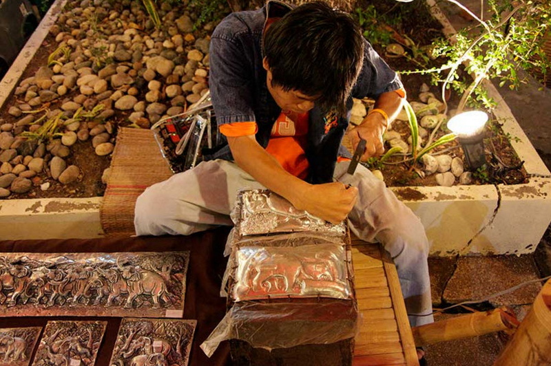Craftsman at Work at Old Chiangmai Cultural Center Person working on metal piece with tools surrounded by plants and rocks at Old Chiangmai Cultural Center.