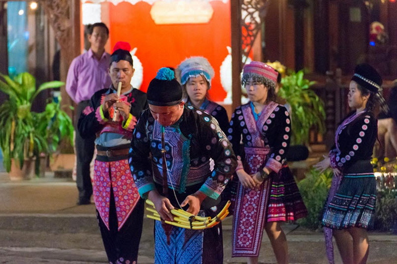 People in traditional attire participating in cultural activity outside a building with plants and a red backdrop at Old Chiangmai Cultural Center Khantoke.