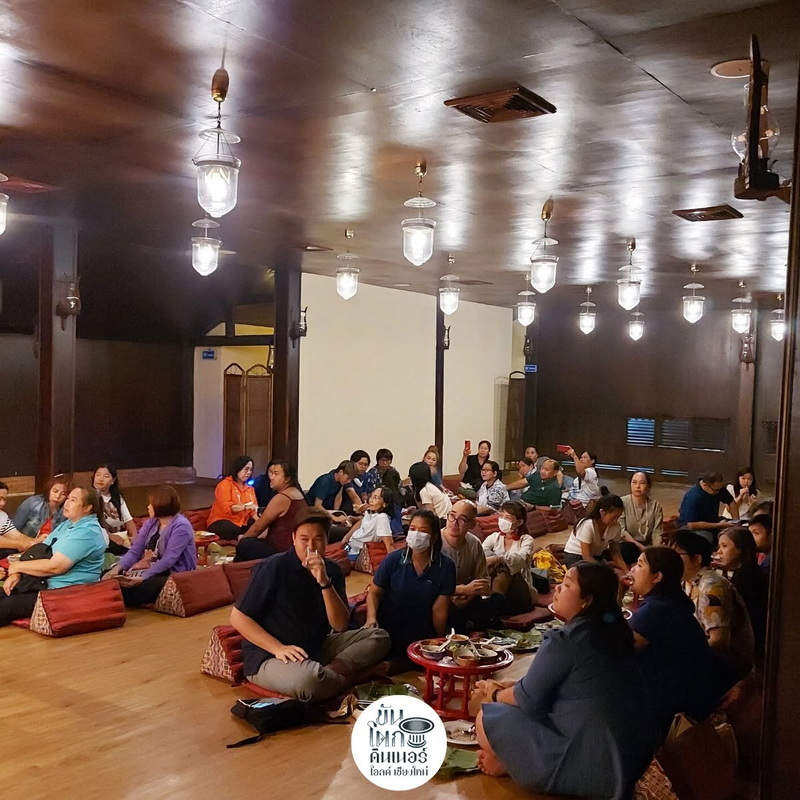 People sitting on the floor inside a wooden hall with hanging lights at Old Chiangmai Cultural Center.