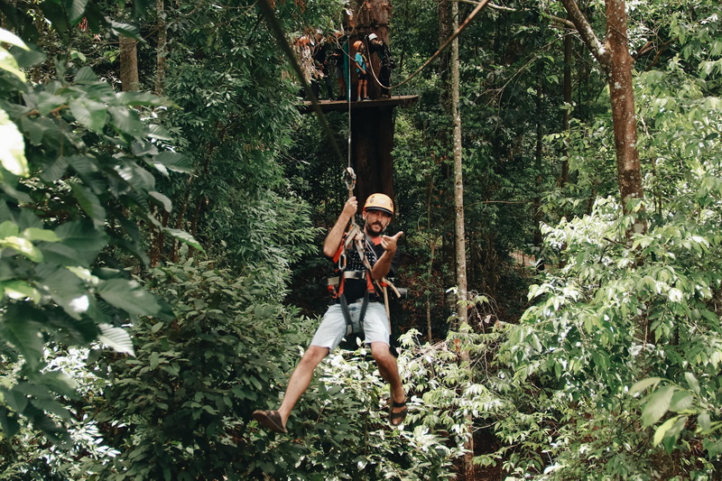 Jungle Adventure Swing at Jungle Flight Zipline Chiang Mai Man swinging on ropes course among lush green trees in forest canopy at Jungle Flight Zipline Chiang Mai.