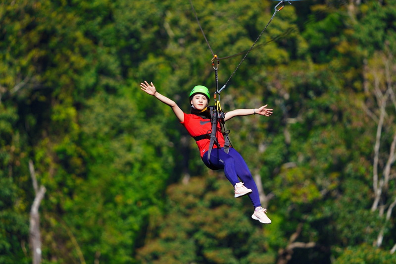 Girl in a red shirt and green helmet on a zipline among green trees at King Kong Smile Zipline.