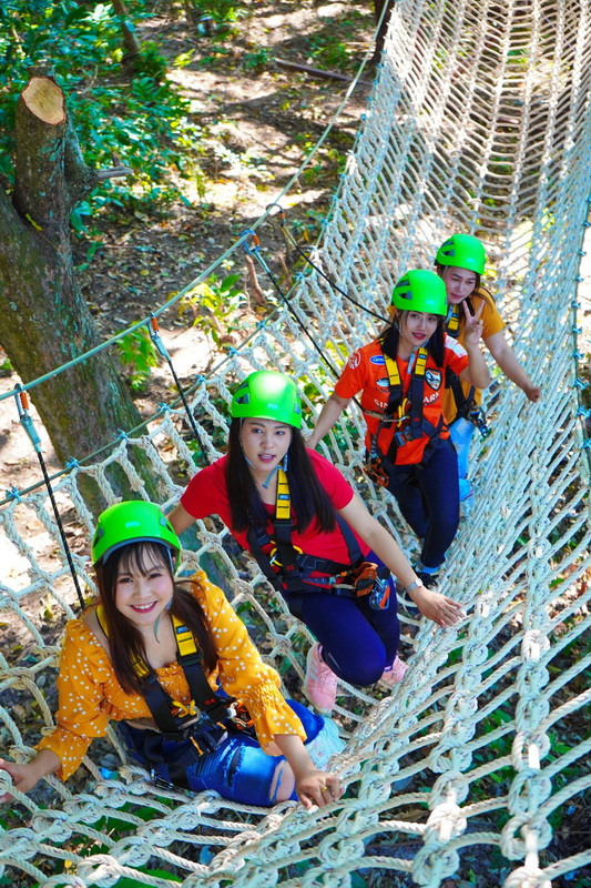 Four women with green helmets and harnesses crossing a rope bridge in dense forest at King Kong Smile Zipline.