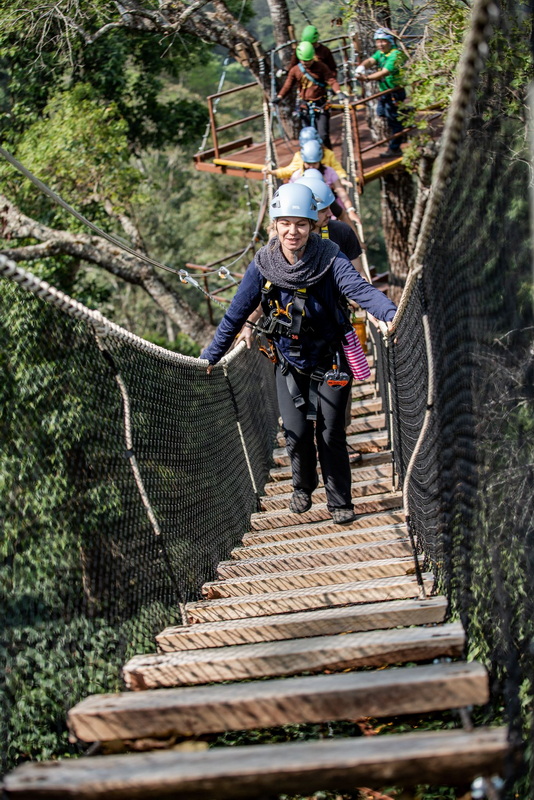 People crossing a wooden suspension bridge through a dense forest canopy at Kingkong Smile Zipline.