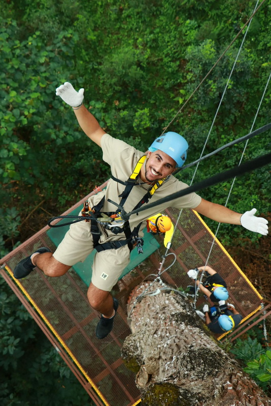 Forest Tree Climber Adventure at Kingkong Smile Zipline Man smiling and hanging from a safety harness in a green forest canopy at Kingkong Smile Zipline.