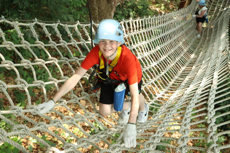 Treetop Rope Climb Adventure at Kingkong Smile Zipline Boy wearing helmet and red shirt climbing a rope net in a wooded area at Kingkong Smile Zipline.