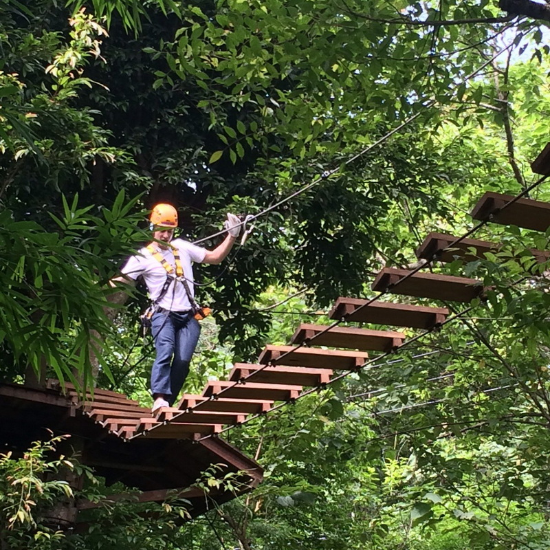 Person in helmet crossing a wooden suspension bridge amidst lush green trees at Phoenix Adventure Park.