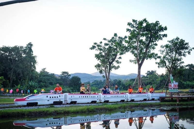 Group of people riding a train-shaped amusement ride near water with trees and mountains nearby at Phoenix Adventure Park.