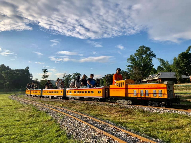 Yellow and orange train with passengers riding outdoors on grassy tracks under clouds at Phoenix Zipline Chiang Mai.