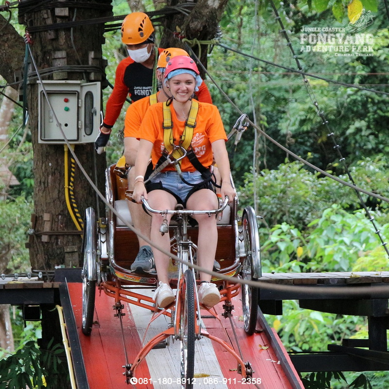 Woman riding a bicycle platform across a wooden bridge in a lush forest at Pong Yang Zipline and Jungle Coaster.