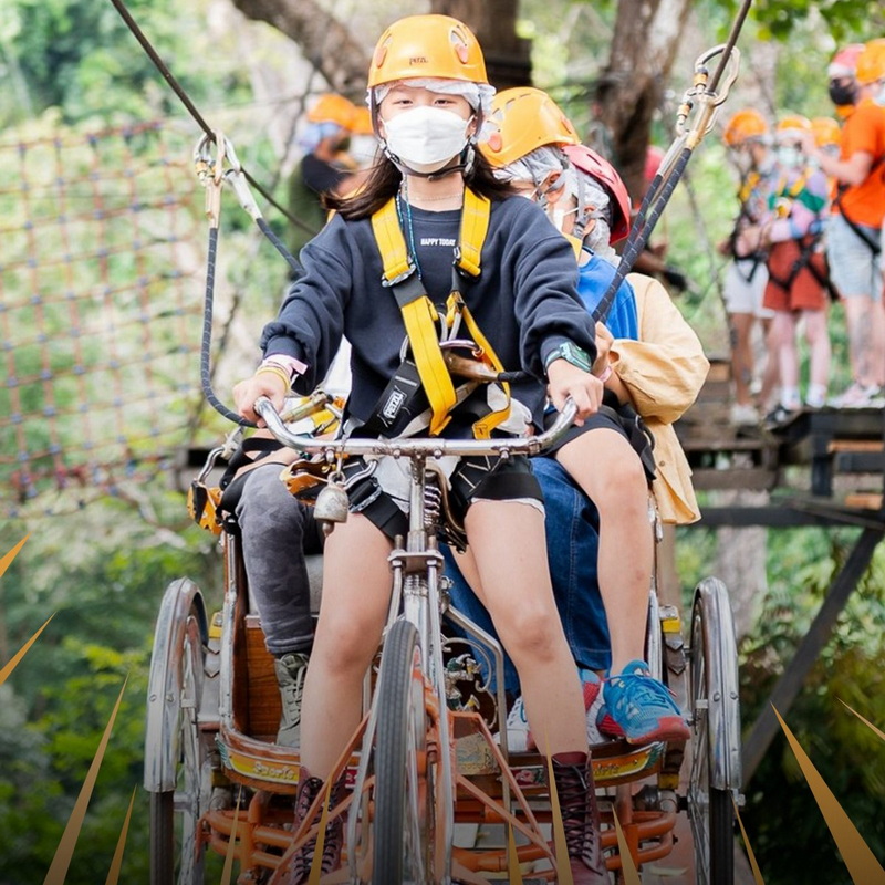 Children wearing helmets and masks riding a rickshaw through forest with other people in background at Pong Yang Zipline and Jungle Coaster.