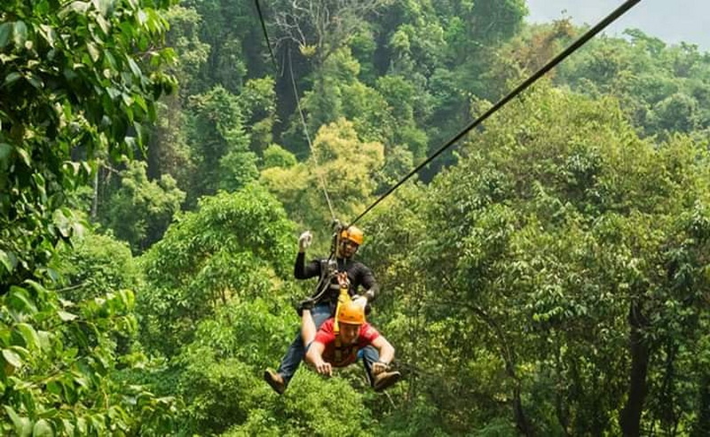 Two people ziplining through lush green trees with safety helmets and harnesses at Pong Yang Zipline.