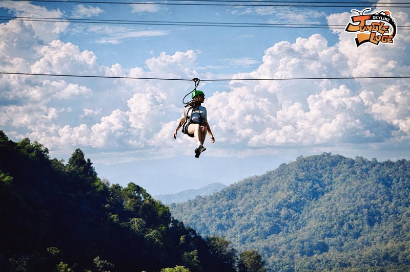 Person riding a zipline over lush mountain forest under blue sky with clouds at Chiang Mai Skyline Adventure.