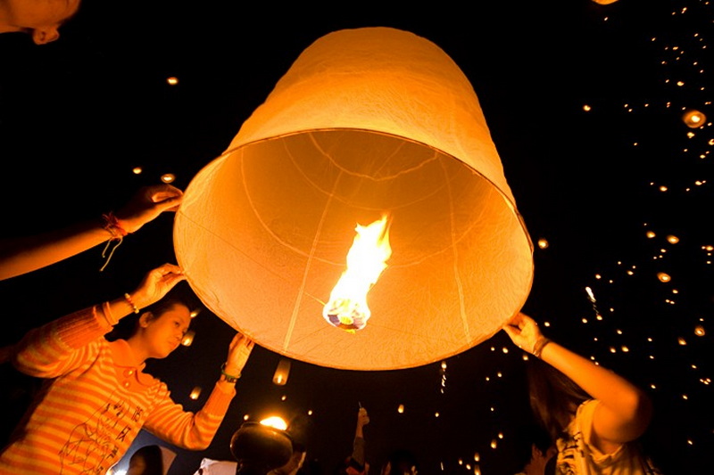 Two individuals release a glowing sky lantern during a night celebration with floating lanterns around at Yi Peng Chiang Mai.