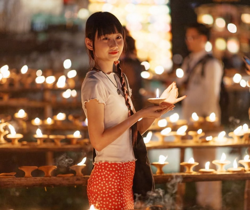 Young woman holding lit candle in a candlelit setting with others around at Yi Peng Chiang Mai.