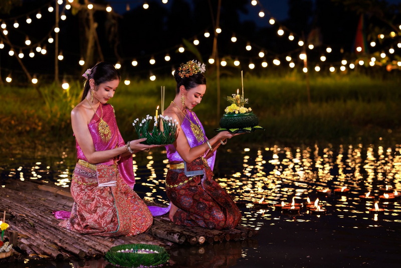 Two women in traditional Thai clothing holding candles by a illuminated water scene at night at Yi Peng Chiang Mai.