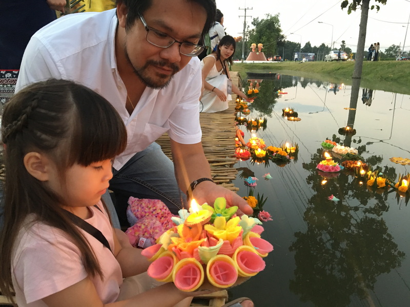 Man and young girl lighting a flower lantern on water with floating lanterns around them at Yi Peng Chiang Mai.