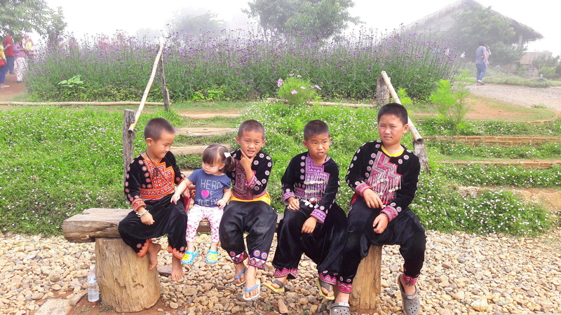 Children dressed in traditional clothes sitting outdoors on wooden logs in a garden setting at Mon Jam.