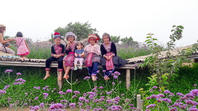 Children dressed in traditional attire sitting on a wooden platform among purple flowers at Mon Jam.