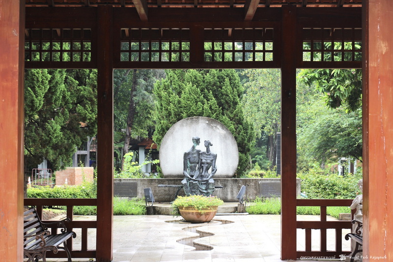 View through wooden pavilion showing a sculpture of two seated figures in a garden with trees at Royal Flora Ratchaphruek.