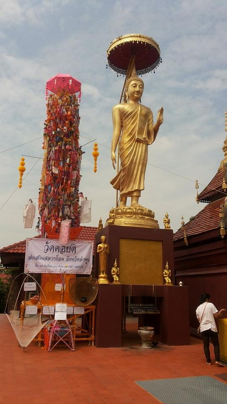 Golden Buddha statue holding an umbrella with prayer flags and temple surroundings at Kruba Sri Wichai Monument.