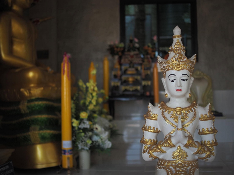 White and gold deity statue with crown and jewelry indoors, with floral arrangements behind at Wat Phra That in Kwaen.