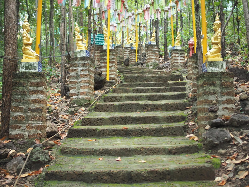 Stone staircase with moss leading through a forest with golden statues and colorful flags at Phra That in Kwaen.