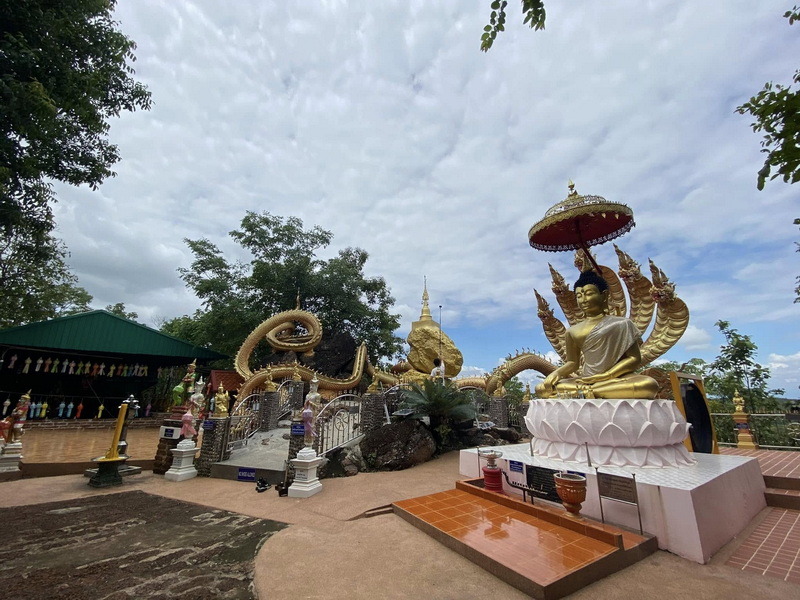 Golden Deity and Serpent Statue at Wat Phra That in Kwaen Golden seated deity statue with serpent and stupa under cloudy sky at Wat Phra That in Kwaen.