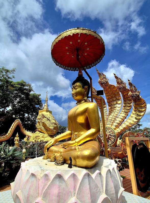 Golden Buddha statue with umbrella and serpent sculpture in outdoor setting during daytime at Phra That in Kwaen.