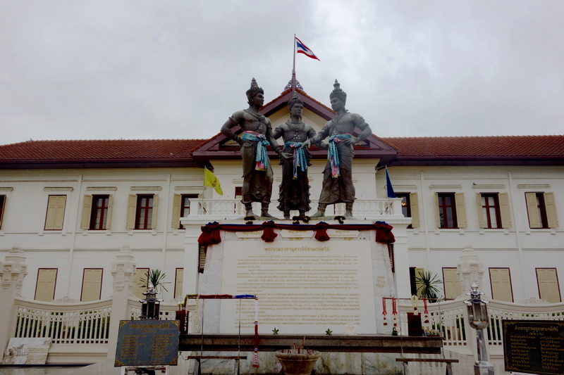 Statues of three figures on a monument in front of a white building with a Thai flag at Three King Monument.