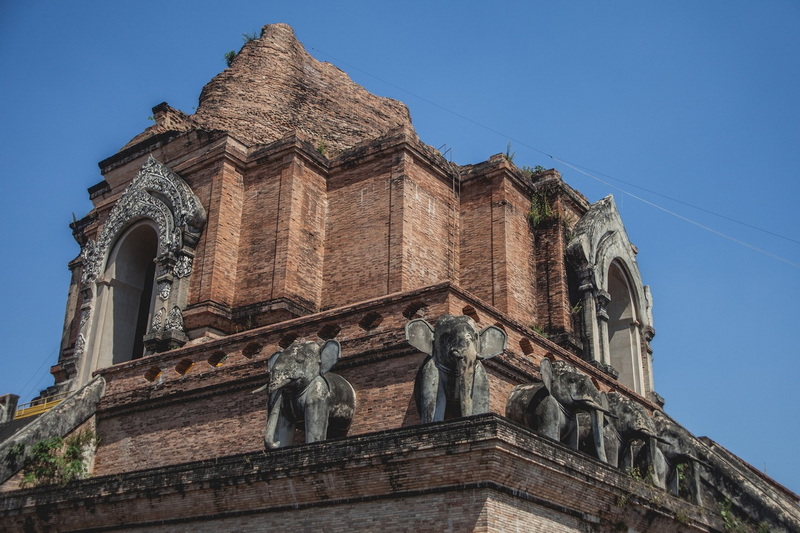 Ancient brick structure with elephant carvings and arched windows under a blue sky at Wat Chedi Luang.