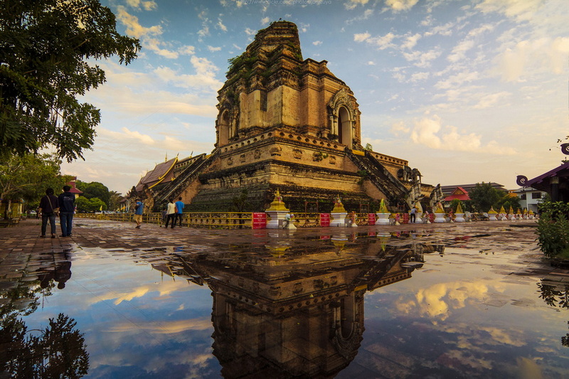 Ancient temple with multiple tiers reflected in a puddle on a cloudy sky day at Chedi Luang temple.