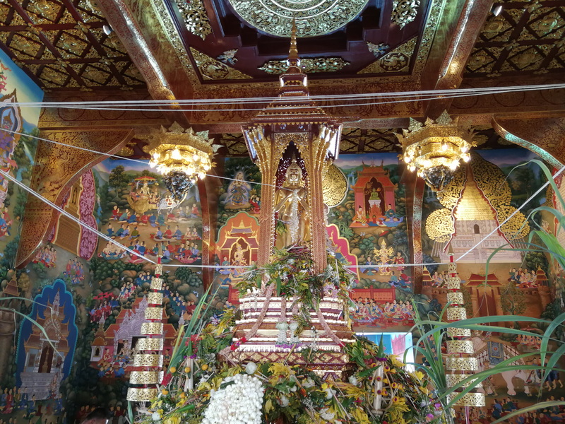 Golden deity statue inside a richly decorated temple with murals and hanging chandeliers at Chedi Luang temple.