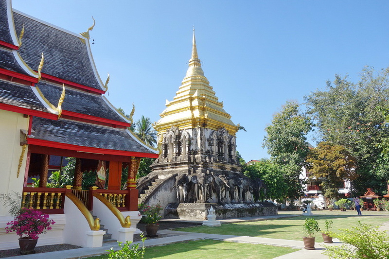 Golden stupa beside temple with decorative roof and surrounding greenery at Wat Chiang Man.