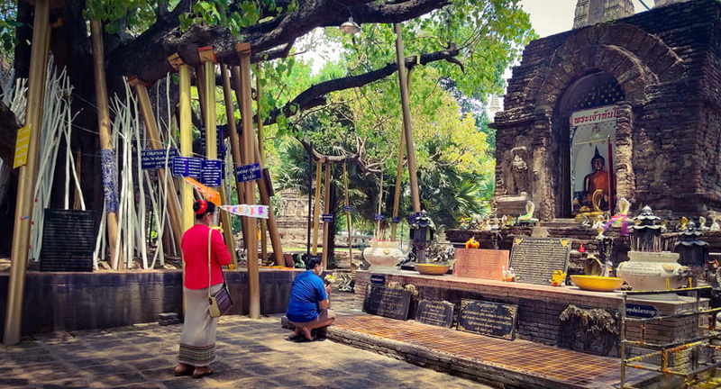 People praying at an outdoor shrine with a Buddha statue amid trees and stone structures at Wat Jed Yod.