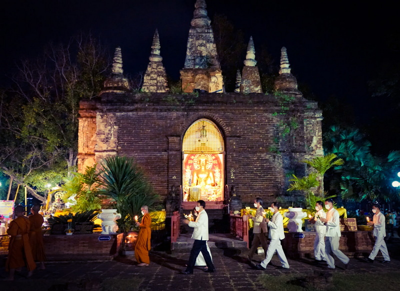 Night scene of a brick stupa with spires illuminated, with monks and visitors walking nearby at Wat Jed Yod.
