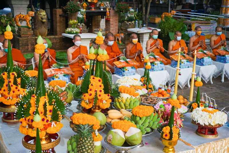 Monks seated behind a decorated table with flowers and fruits during a ceremony at Wat Jed Yod.